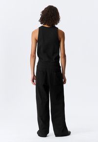 Άτομο με σγουρά μαλλιά, που στέκεται πλάτη, φοράει μαύρο αμάνικο τοπ και φαρδιά μαύρη παντελόνια με τσέπες στο πίσω μέρος.