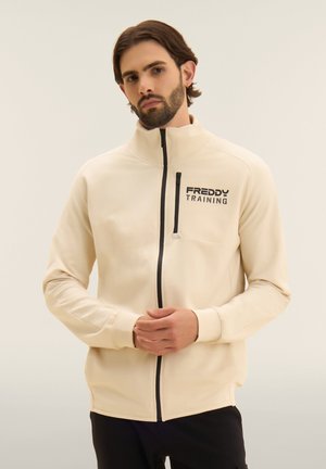 Parrakas mies, joka käyttää kermaista vetoketjullista takkia, jossa on "FREDDY TRAINING" -logo, seisoo kätensä edessä ristissä neutraalia taustaa vasten.