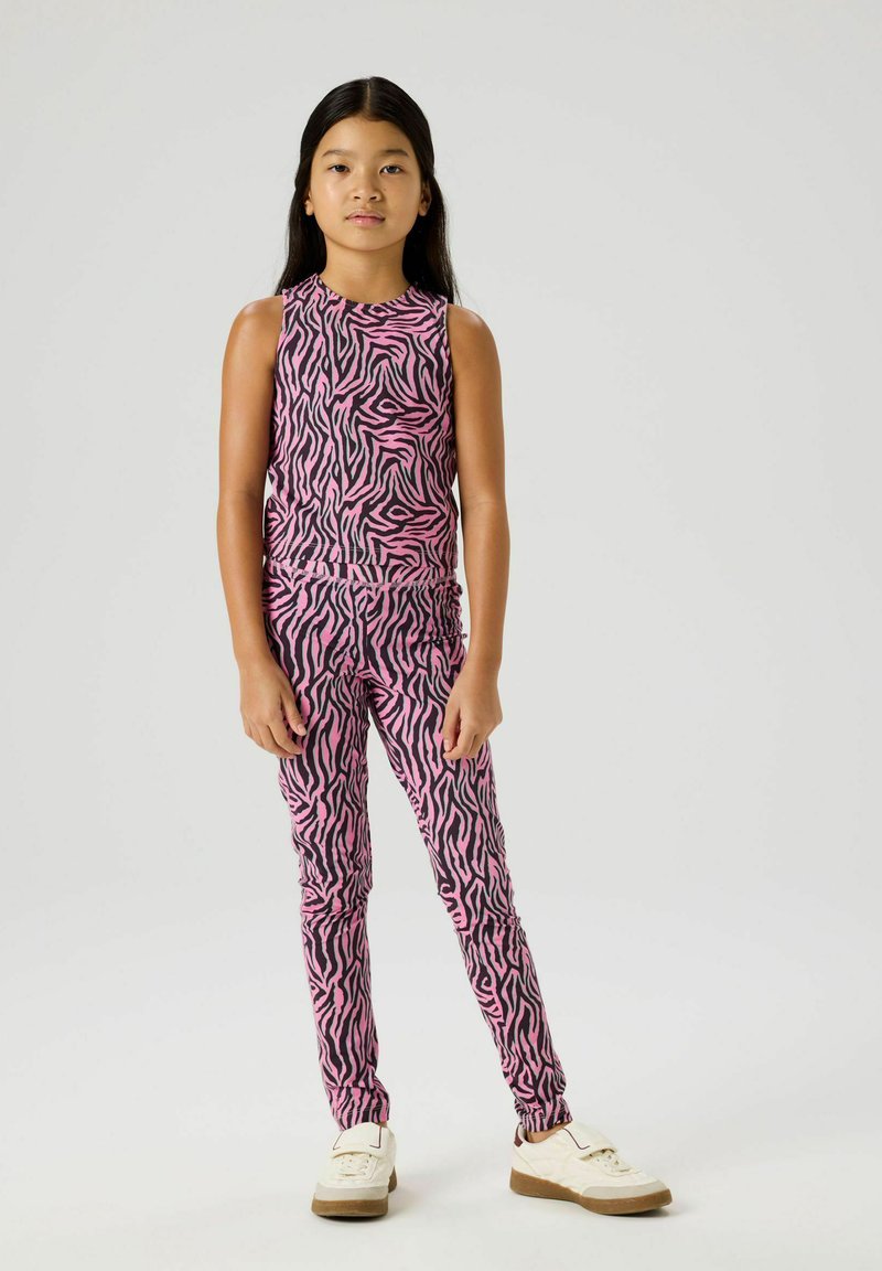 Meisje staand in een mouwloze top en legging met roze en zwarte zebra-print, met witte sneakers, tegen een effen lichte achtergrond.