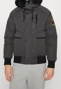 Herre jakke i mørkegrå puffer med en kunstpelshætte, lynlås- og klaplommer, elastiske ærmer og et mærke på ærmet.