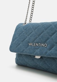 Quiltad denimhandväska i blått med en silver kedjestraps och logodetaljer. Har blindknäppning och strukturerad mönster.