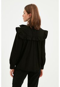 Blusa nera con colletto arricciato, maniche lunghe e dettaglio dei bottoni sul retro. Realizzata in tessuto leggero, presenta una vestibilità ampia.