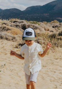 Niño vestido con una camisa de botones beige claro y pantalones cortos grises claros, con una gorra en blanco y negro que tiene un logo, caminando sobre un terreno arenoso.