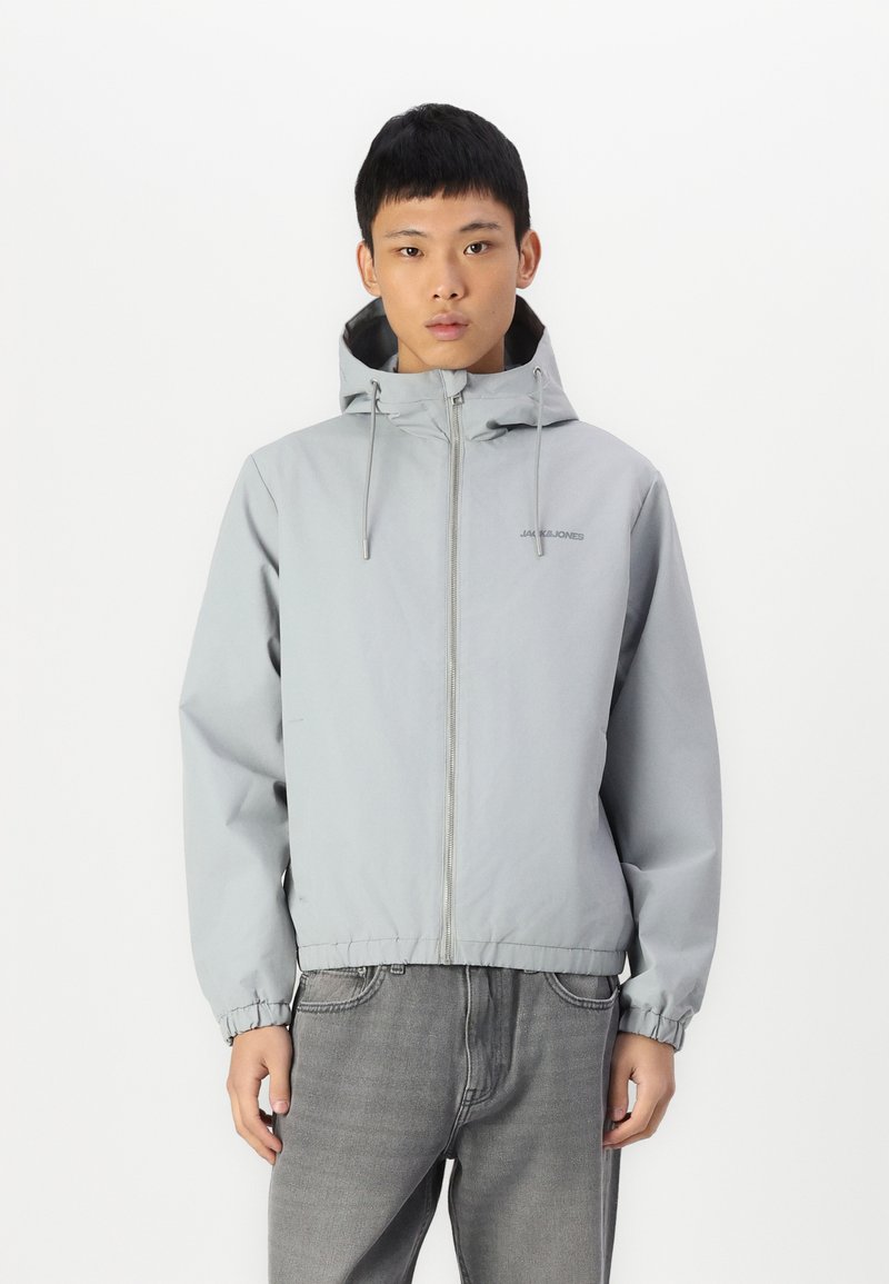 Grijze windbreaker jas met een capuchon, rits aan de voorkant, elastische manchetten en een rechte zoom. Heeft een subtiele logo op de borst.