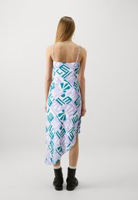 Asymmetrisk kjole i pastell lilla stoff med dristige teal og hvite geometriske mønstre, med tynne stropper og en flytende silhuett.