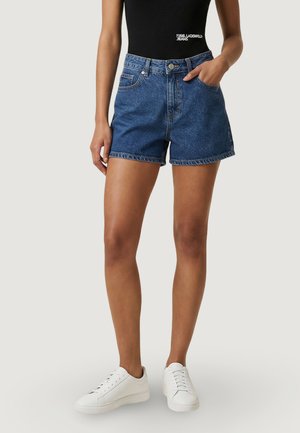 Donna con pantaloncini di jeans blu medio, top nero senza maniche con logo e sneaker bianche, in piedi davanti a uno sfondo chiaro e uniforme.