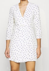 Robe enveloppante blanche à manches trois-quarts, ornée de pois bleu marine et dotée d'une taille à nouer. Tissu doux avec un décolleté en V profond.