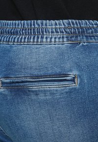 Κοντινό πλάνο σε μπλε τζιν παντελόνια με ελαστική μέση και οριζόντια πίσω τσέπη ραμμένη σε σκοτεινό υφασμάτινο φόντο.