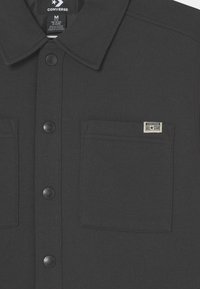 Sort skjorte med knapper foran, krave, to brystlommer, glat tekstur og en lille "Converse All Star" logo-tag.