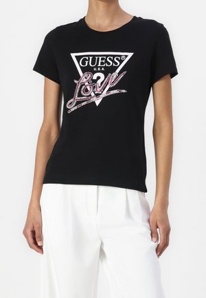 Czarny t-shirt z krótkim rękawem z białym trójkątnym logo z napisem "GUESS U.S.A." oraz różowym kryształkowym napisem "Love" pisaną kursywą, noszony z białymi spodniami.