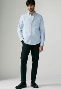 Chemise boutonnée rayée bleu clair et blanche, associée à un pantalon noir et des chaussures noires, portée par un homme dans une posture décontractée.