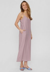 Lungo vestito lavanda con spalline sottili, scollatura a V profonda, tasche laterali e vestibilità comoda. Abbinato a sandali con tacco a blocco di color blu chiaro.