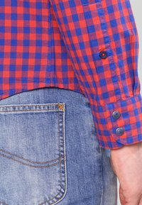 Ärmel eines rot-blau karierten Hemdes mit dunklen Knöpfen neben der Gesäßtasche einer hellblauen Jeans und der sichtbaren Hand einer Person.