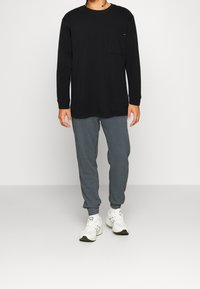 Uomo in camicia nera a maniche lunghe, pantaloni jogger grigi e sneakers bianche in piedi davanti a uno sfondo chiaro e uniforme.