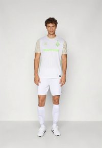 Λευκή φανέλα ποδοσφαίρου με ανοιχτό μπεζ μανίκια, με πράσινες λεπτομέρειες και κάθετες ρίγες. Συνδυάζεται με λευκό σορτς και κάλτσες.