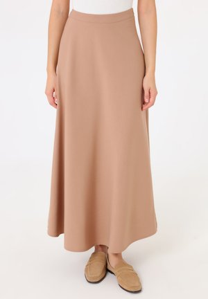 Donna che indossa una lunga gonna beige a linea ad A e mocassini beige coordinati, in piedi con le mani rilassate lungo i fianchi su sfondo bianco.