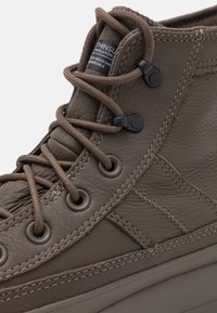 Primer plano de bota alta de cuero marrón con cordones gruesos, ojales de metal, lengüeta acolchada y detalles de costura visibles.