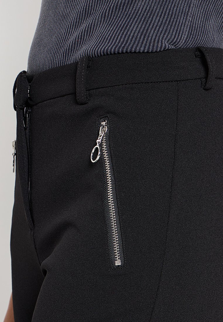 Pantalons noirs avec une texture lisse, coupe ajustée et deux poches zippées avec quincaillerie en argent à l'avant.