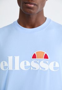 Gaiši zils kokvilnas T-krekls ar lieliem baltajiem burtiem un izšūtu logotipu ar sarkaniem, oranžiem un krēma akcentiem virs teksta.