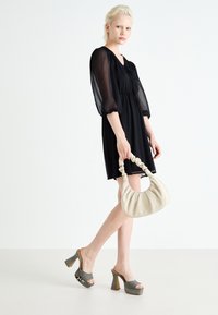 Robe noire courte avec manches bouffantes, taille cintrée, associée à des mules à plateforme grises et un sac à main crème avec une anse torsadée.
