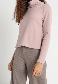 Jasnoróżowy sweter z golfem o ribbowanym wzorze, z długimi rękawami i lekko skróconym krojem, połączony z brązowymi spodniami w kratkę o wysokim stanie.