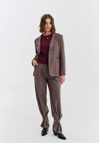 Žena v hnedom šitom obleku s nariasenými detailmi na gombíky pri členku, v kombinácii s bordovým vrškom a čiernymi čižmami na podpätku.