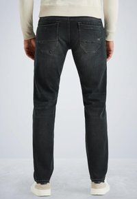 Muž oblečený v čiernych džínoch a béžovej mikine, stojí so uvoľnenými rukami popri tele, viditeľný zozadu na jednotnom pozadí.