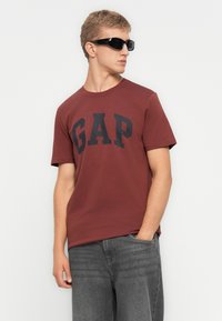 Ung mand iført sorte solbriller, en maroon GAP t-shirt og grå jeans står med den ene hånd i lommen og kigger til siden.