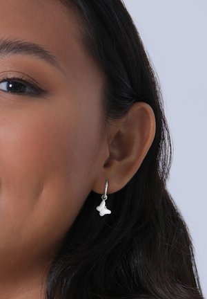 Weiße schmetterlingsförmige Ohrringe, die an einem silbernen Ring hängen und am Ohr eines Models präsentiert werden. Glatte, polierte Oberfläche mit subtilen Details.