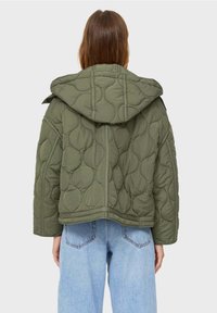 Olivgrüne gesteppte Jacke mit Kapuze, mit einem kastigen Design und auffälligen Steppmuster. Getragen über hellblauen Jeans.