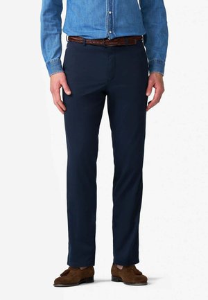 Uomo con pantaloni blu scuro, cintura intrecciata marrone, camicia di jeans blu e mocassini marroni con nappine, in piedi su sfondo bianco.