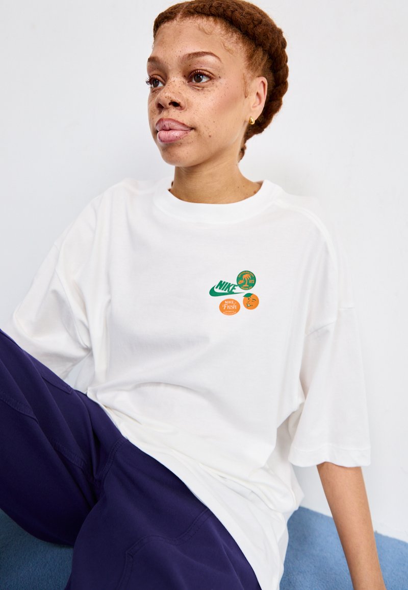 Camiseta blanca de gran tamaño hecha de algodón, con logotipos gráficos en verde y naranja en el pecho. Manga corta, escote redondo, textura suave.