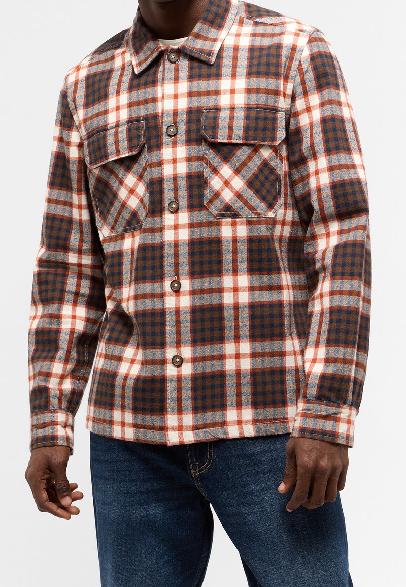 Károvaná flanelová košile se vzorem v hnědé, modré a červené barvě, se dvěma kapsami na prsou a hnědými knoflíky. Natažená přes modré džínové kalhoty.