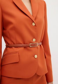Oranje blazer met een getailleerde pasvorm, gouden knopen en een bruine leren riem. Soepele stoftextuur, ronde revers en details van zijzakken.