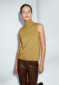 Maglione senza maniche color giallo senape con collo alto, texture a coste, abbinato a pantaloni in pelle marrone. Maglia liscia, silhouette aderente, design minimalista.