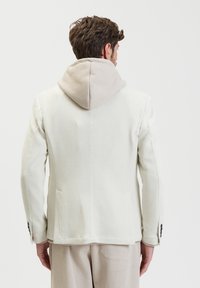 Giacca di tessuto crema con cappuccio a costine beige, due tasche laterali e dettagli con bottoni sulle maniche. Vestibilità su misura, colori e materiali a contrasto.