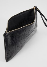 Mustan krokotiilitaskumalli, jossa on kullanvärinen vetoketju, sisäinen tasku ja rannetuki helppoa kantamista varten.