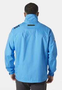 Chaqueta impermeable azul hecha de material suave, con alto cuello, logo negro contraste en el hombro y un detalle vertical en la parte posterior.