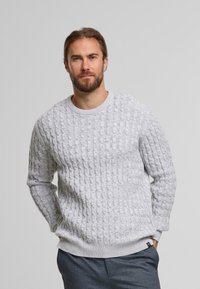Suéter de punto gris claro con un diseño de cable texturizado, cuello redondo y puños de canalé. Llevado sobre pantalones azul marino.