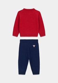 Czerwony sweter z dzianiny z prążkowanym kołnierzem, noszony z granatowymi spodniami dzianinowymi z małą naszywką z logo. Całość wykonana z miękkiego, strukturalnego materiału.