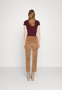 Vrouw met lang haar, gekleed in een bordeauxkleurig shirt met korte mouwen, tan kleurige corduroy broeken en beige hielen, staand tegenover een lichtgrijze muur.