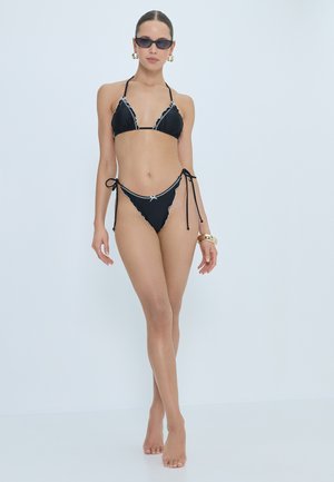 Woman wearing black bikini with white trim, dark sunglasses, gold hoop earrings, and bracelets, standing against a plain light background.