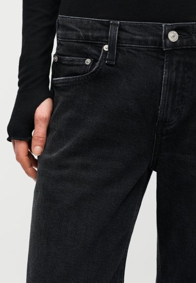 Κοντινό πλάνο μαύρου τζιν παντελονιού με ασημένιο κουμπί και νίκελ κουμπώματα, φορεμένο με μαύρο μακρυμάνικο μπλουζάκι, εμφανίζοντας ένα χέρι που ακουμπάει δίπλα στο μηρό.