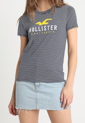 Femme portant un t-shirt rayé noir et blanc Hollister California et une mini-jupe en denim bleu clair avec des bords effilochés.