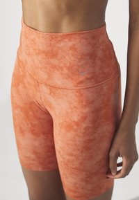 Buste et main féminins portant un short cycliste Nike taille haute orange tie-dye avec un petit logo swoosh sur la hanche gauche.