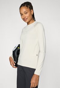 Haut long beige clair à manches longues avec un col rond, texture douce, détail de fermeture éclair sur le côté, porté avec un casque noir sur le côté gauche.