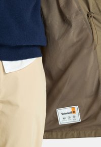 Veste marron avec doublure en mesh, arborant une étiquette Timberland. Portée sur un pull bleu marine et un pantalon beige clair. Détails de couture visibles clairement.