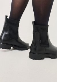 Bottines noires en cuir avec un empiècement en tissu élastique à côtes, dotées d'une semelle en caoutchouc épaisse et d'un logo sur le côté.