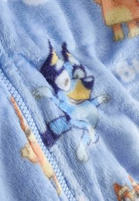 Tissu en polaire bleu doux avec un personnage de chien dessin animé, une fermeture éclair bleu clair, et de petits motifs d'animaux orange sur un vêtement pour enfant.