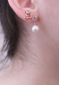 Orecchini in oro rosa con un design a fiocco, impreziositi da una piccola gemma e una perla pendente. La perla è rotonda, liscia e bianca.